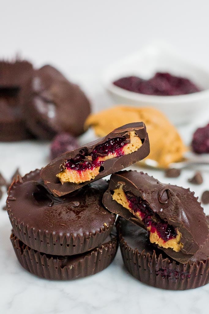 A stack of chocolate berry chia jam peanut butter cups. Two are cut open so you can see the layers. There is a spoonful of peanut butter, a bowl of chia seed jam and more chocolate cups in the background.