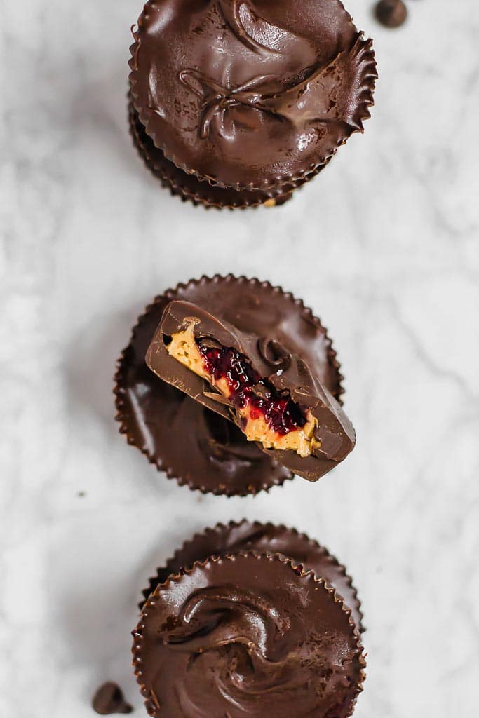 Three chocolate chia jam peanut butter cups in a vertical line sitting on a marble table. The middle chocolate cup is cut in half so you can see the peanut butter and red berry chia jam layers.