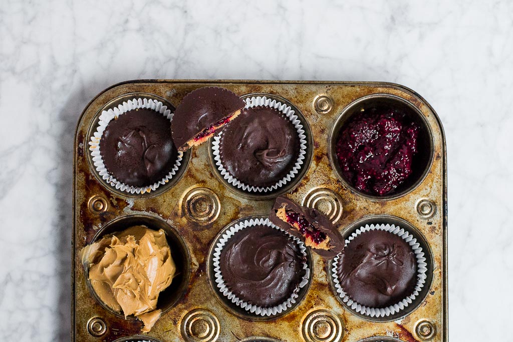 A muffin tin filled with chocolate peanut butter cups. There is one muffin well filled with a red berry chia jam. Another muffin well is filled with creamy peanut butter. There are half segments of chocolate peanut butter cups sitting on top of the muffin tin so you can see the layers inside.