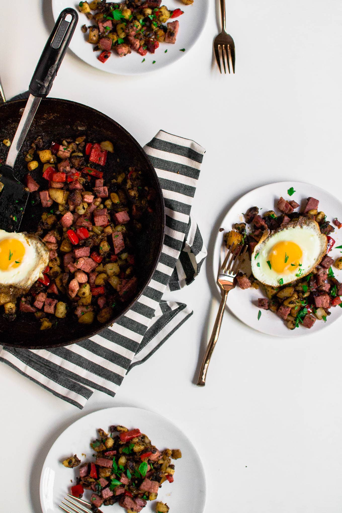 A table set for three with corned beef hash on each plate and a large skillet in the middle holding the leftover corned beef hash. The table is white with a white and grey napkin.
