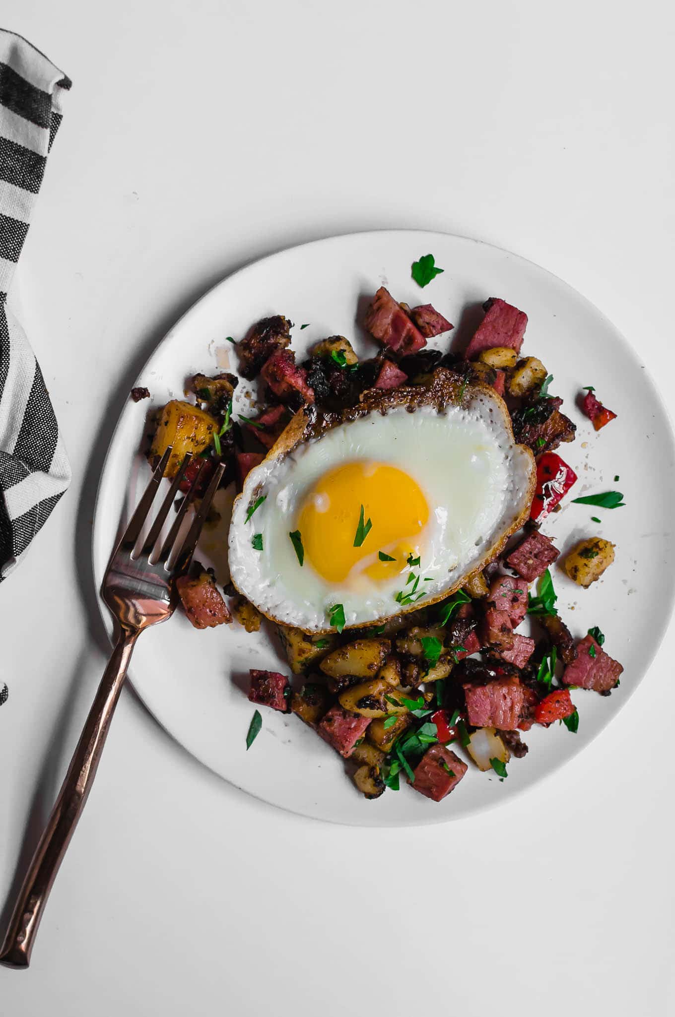 An aerial view of a white plate of gluten-free leftover corned beef hash topped with a sunny side up egg sitting on a white table, with a fork and a gray and white napkin underneath it.