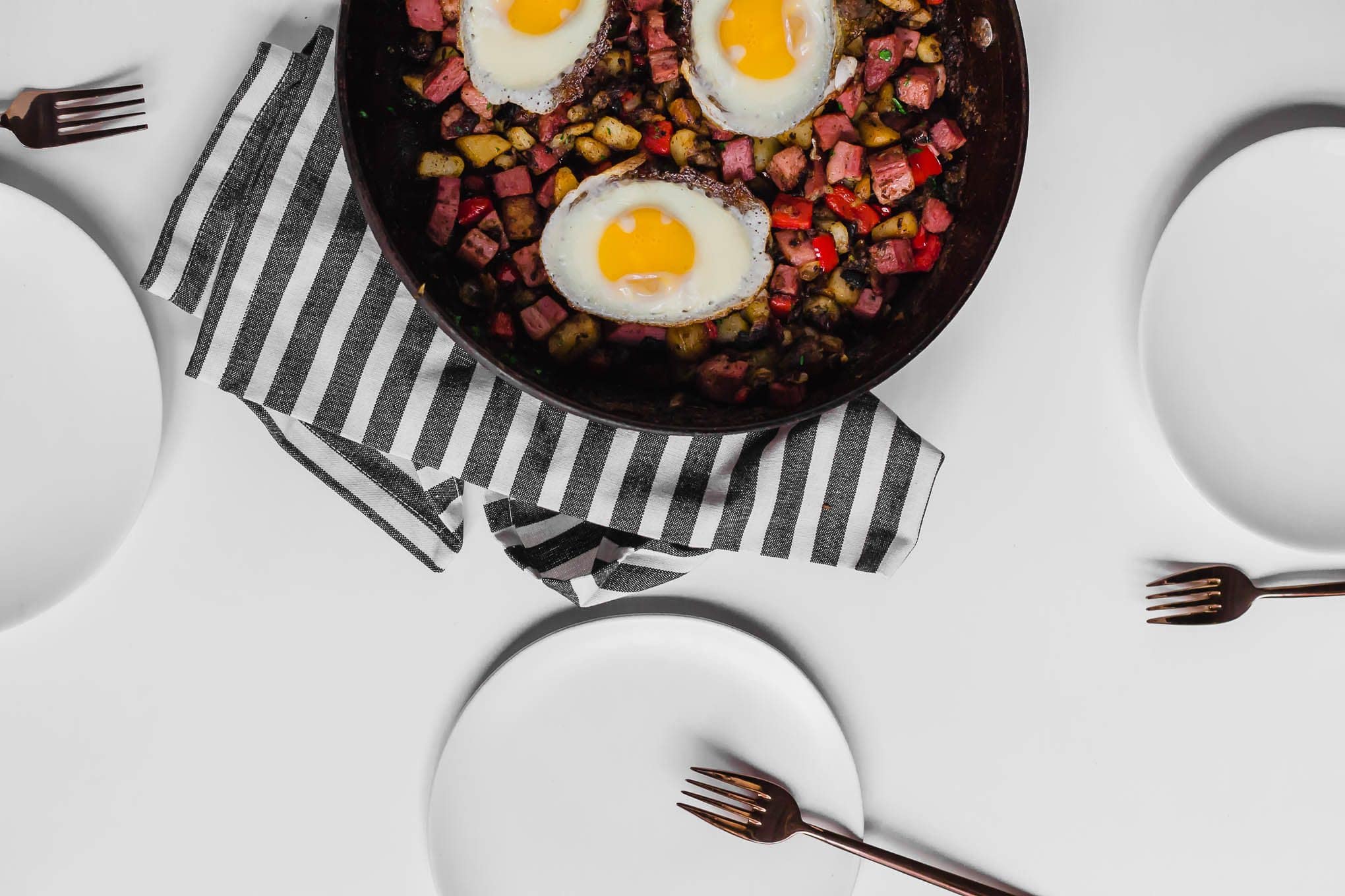 A table set for three with corned beef hash on each plate and a large skillet in the middle holding the leftover corned beef hash. The table is white with a white and grey napkin.