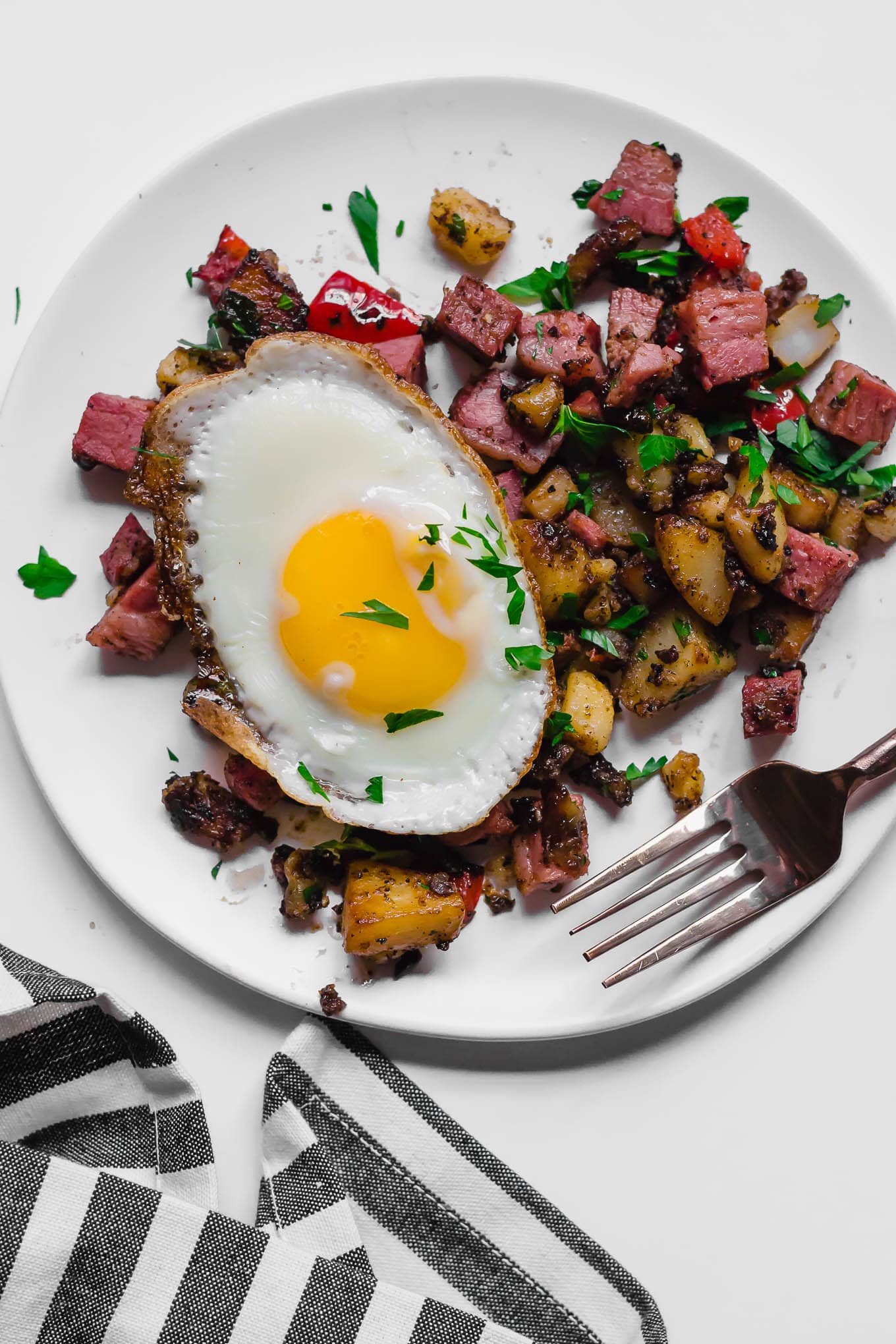 An aerial view of a white plate of gluten-free leftover corned beef hash topped with a sunny side up egg sitting on a white table, with a fork and a gray and white napkin underneath it.