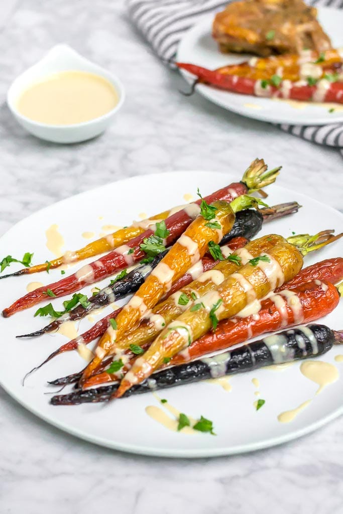 A plate of Roasted Rainbow Carrots with Lemon Tahini Sauce is drizzled with creamy sauce and garnished with chopped herbs. A bowl of extra sauce and a dish with food appear in the background on a marble surface.