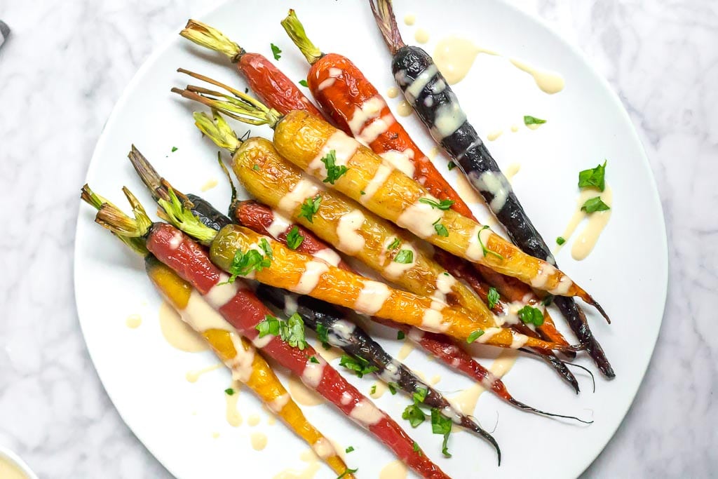 A plate of Roasted Rainbow Carrots with Lemon Tahini Sauce, garnished with chopped fresh herbs, is served on a white plate atop a marble surface.
