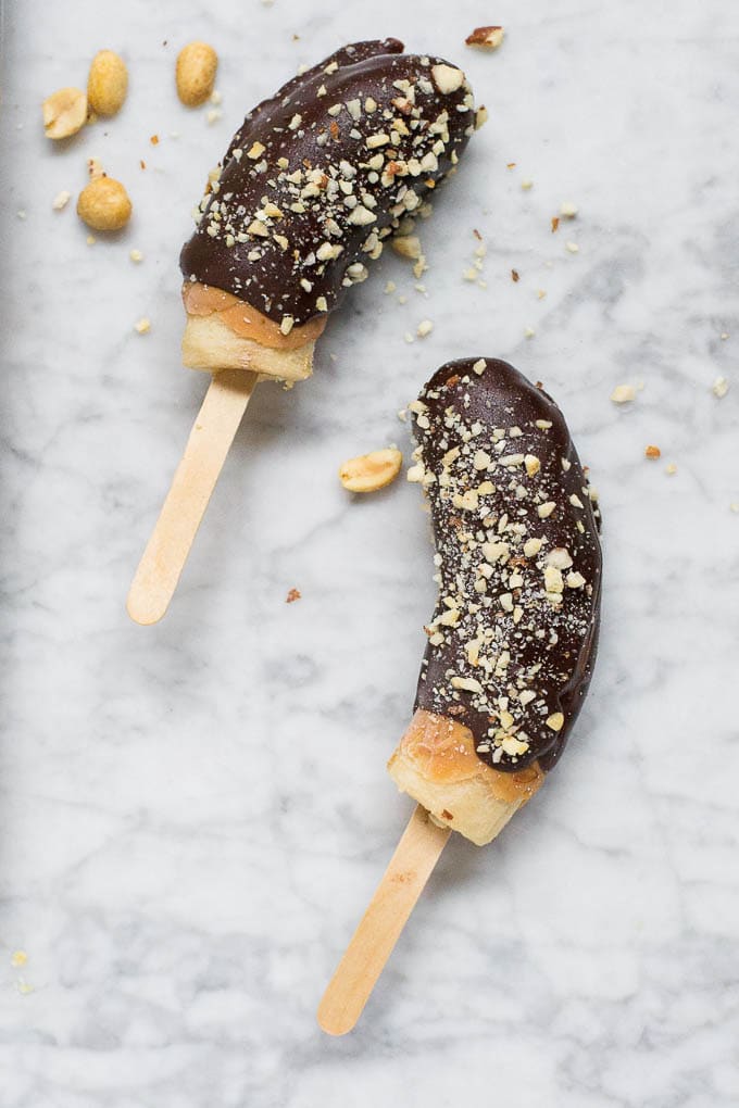 Two half bananas on wooden sticks are transformed into Chocolate Frozen Banana treats, coated in chocolate and sprinkled with chopped peanuts, resting on a light marble surface with a few peanut pieces scattered around.