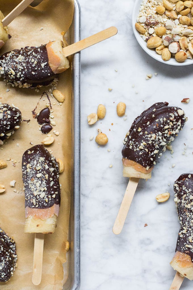 Chocolate Frozen Bananas on wooden sticks are dipped in rich chocolate and sprinkled with chopped nuts, arranged on parchment paper over a marble surface, with a small bowl of mixed nuts nearby.