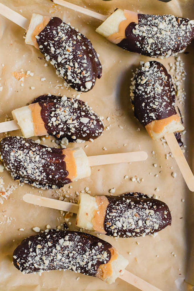 Chocolate Frozen Banana halves on wooden sticks, partially dipped in chocolate and sprinkled with chopped nuts, arranged on parchment paper.