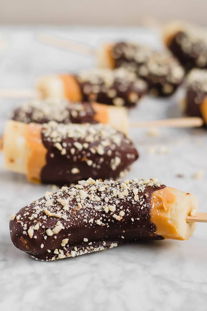 Close-up of chocolate frozen banana halves on sticks, partially dipped in chocolate and sprinkled with chopped nuts, arranged on a light marble surface.