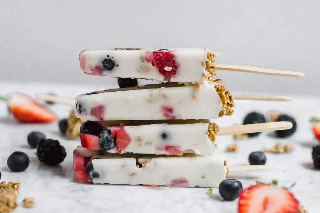 Four Greek yogurt popsicles with pieces of berries and granola are stacked on a white surface, surrounded by scattered fresh berries and crunchy granola pieces.