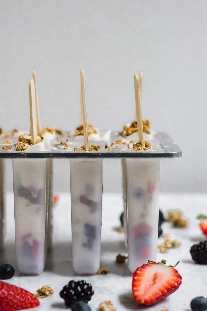 A popsicle mold filled with creamy greek yogurt popsicles and mixed berries, topped with granola and wooden sticks. Fresh strawberries, blackberries, and granola pieces are scattered around the mold on a light surface.
