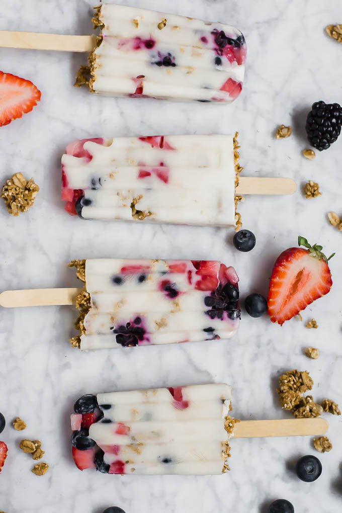 Four Greek yogurt popsicles with granola and mixed berries are arranged on a marble surface, surrounded by scattered strawberries, blueberries, blackberries, and crunchy granola clusters.
