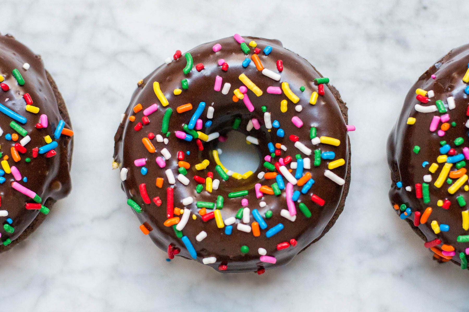 An aerial view of three gluten-free vegan baked chocolate donuts with chocolate glaze and rainbow sprinkles.