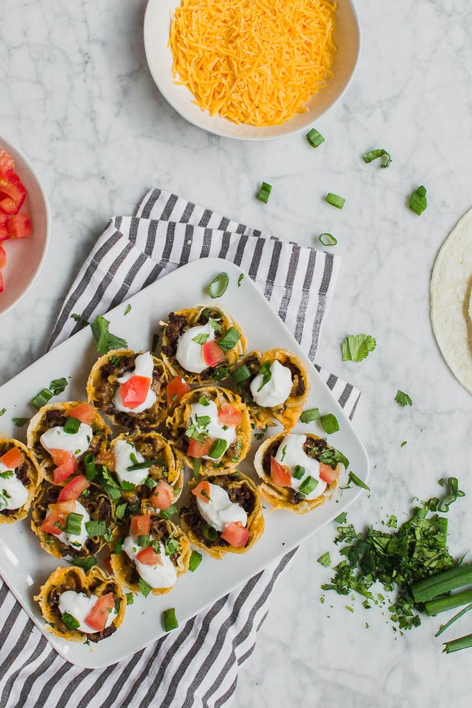 An aerial view of mini beef taco cups on a white plate, surrounded by bowls of toppings - tomatoes, cheddar cheese, green onions, cilantro and some corn tortillas on a white marble table.