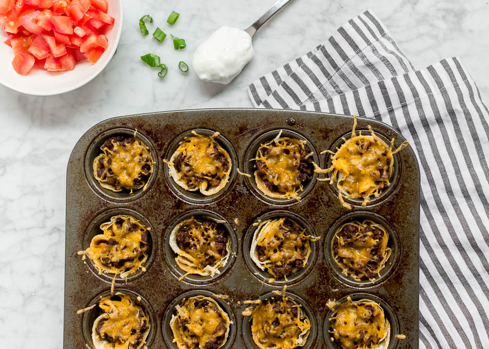 A mini muffin tin filled with corn tortillas, ground beef and cheddar cheese fresh from the oven. Surrounded by tomatoes, green onion and sour cream toppings.