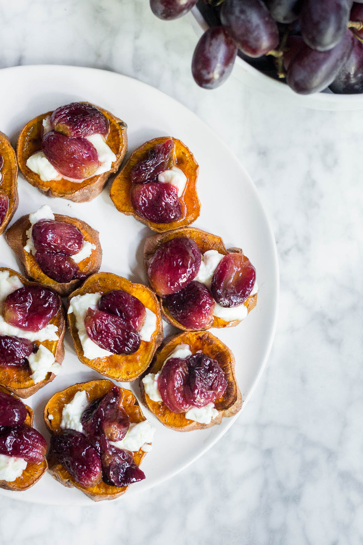 Aerial view of a plate of sweet potato rounds with roasted grapes, goat cheese and honey and a bowl of fresh grapes