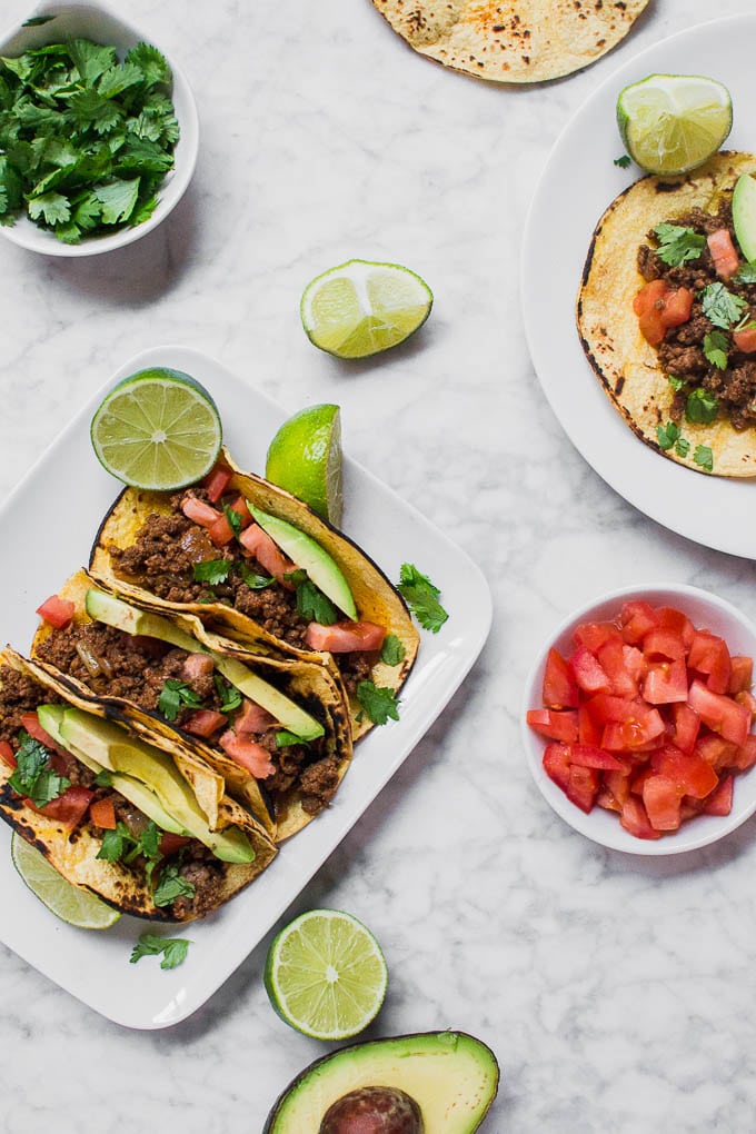An aerial view of a plate of gluten-free ground beef tacos, with a bowl of diced tomatoes, a half of an avocado, a bowl of cilantro, and limes.