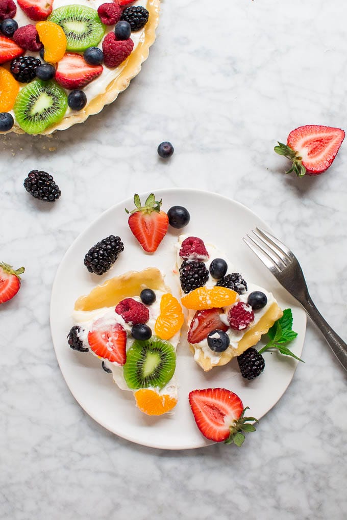 Aerial view of a white plate with two slices of fruit tart toped with mandarin orange, kiwi, and berries with a fork and a large tart in the upper left corner.