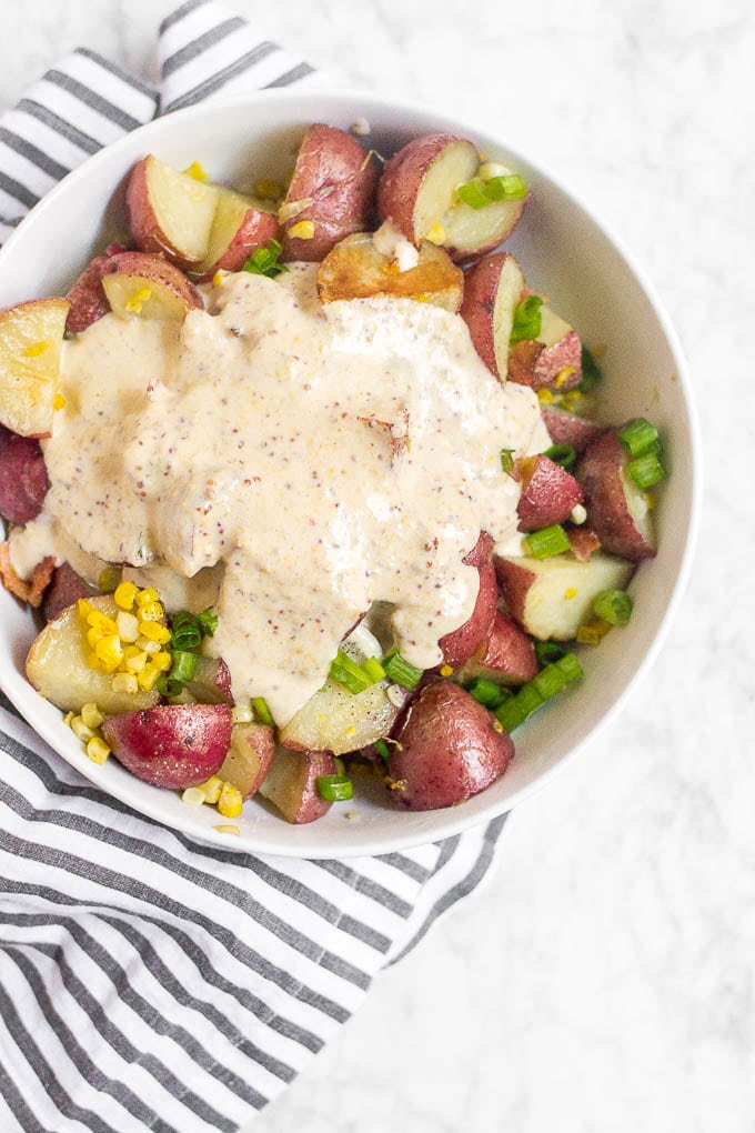 A white bowl filled with Roasted Corn and Bacon Red Potato Salad, featuring roasted red potatoes, chopped green onions, and roasted corn, all topped with a creamy mustard sauce and set on a striped cloth over a light marble surface.