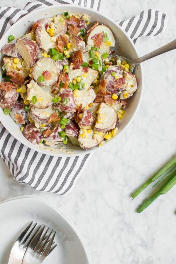 A bowl of Roasted Corn and Bacon Red Potato Salad with sliced red potatoes, corn, bacon bits, and chopped green onions sits on a striped cloth. A serving spoon rests in the bowl, with a plate and forks nearby.