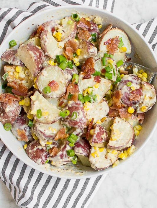 A bowl of Roasted Corn and Bacon Red Potato Salad with sliced red potatoes, corn, bacon bits, and chopped green onions sits on a striped cloth. A serving spoon rests in the bowl, with a plate and forks nearby.