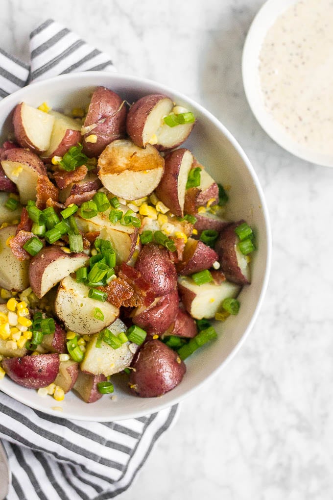 A bowl of Roasted Corn and Bacon Red Potato Salad with chopped green onions sits on a striped cloth, next to a small dish of creamy dressing, all arranged on a marble surface.