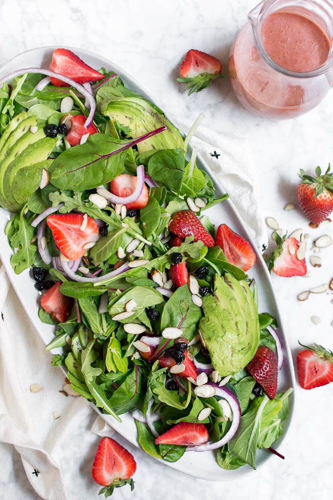 An aerial view of a platter with spinach salad and strawberries, red onion, slivered almonds, dried blueberries and a strawberry balsamic dressing.