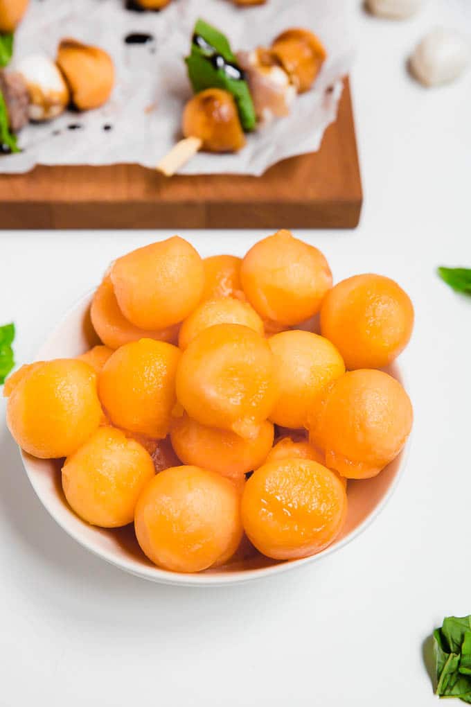 A bowl of cantaloupe balls with cantaloupe mozzarella prosciutto skewers in the background