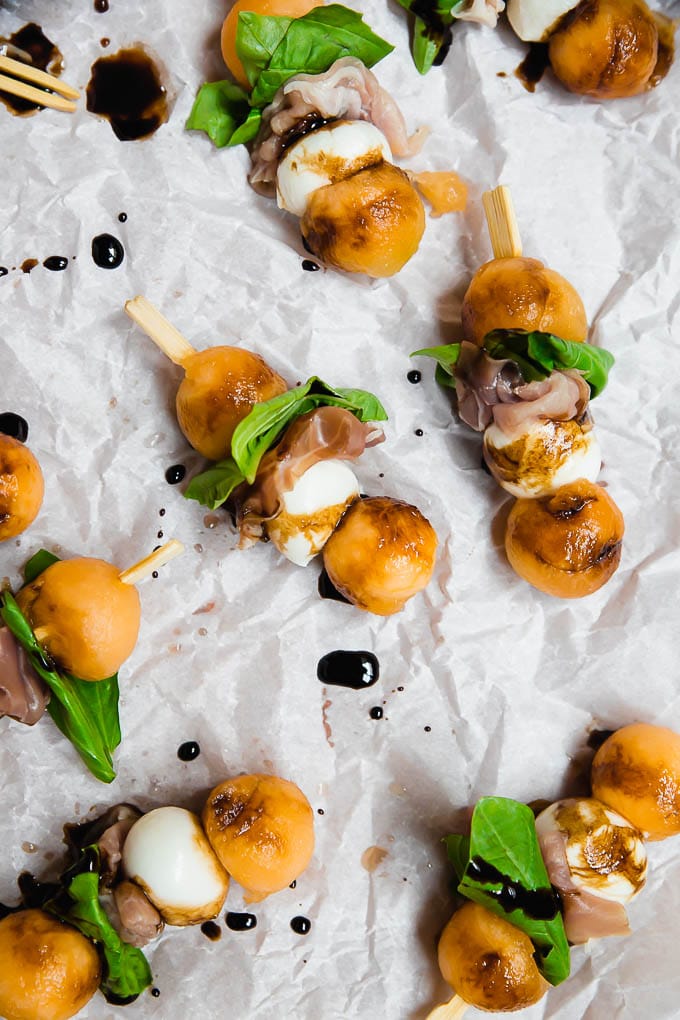 Aerial view of melon mozzarella balls prosciutto and basil on an appetizer skewer drizzled with balsamic vinegar glaze on white parchment paper