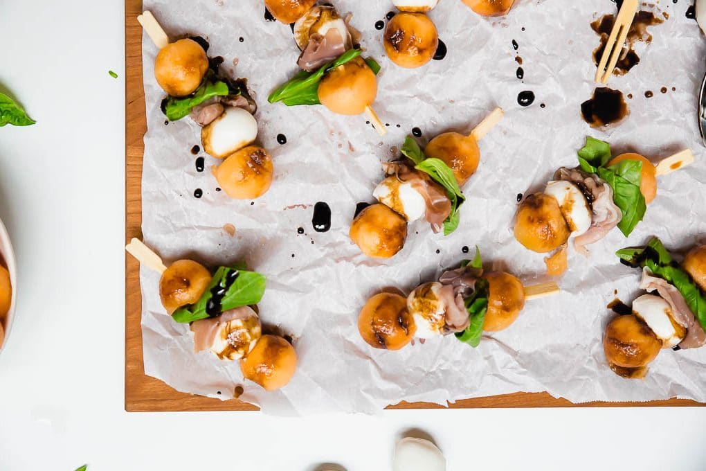 A serving board with mini skewers of melon mozzarella prosciutto and basil drizzled with balsamic vinegar glaze.