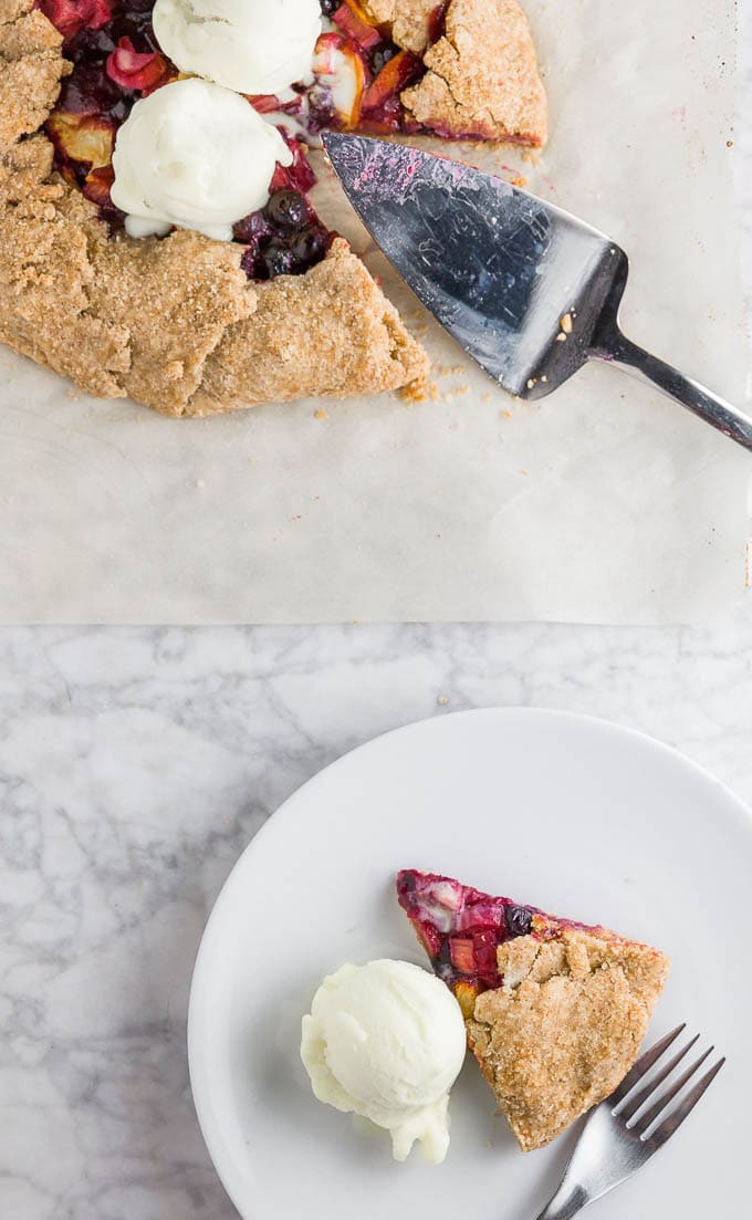 A gluten-free peach blueberry rhubarb galette on a marble table and a piece of galette on a white plate with a scoop of vanilla ice cream and a fork.