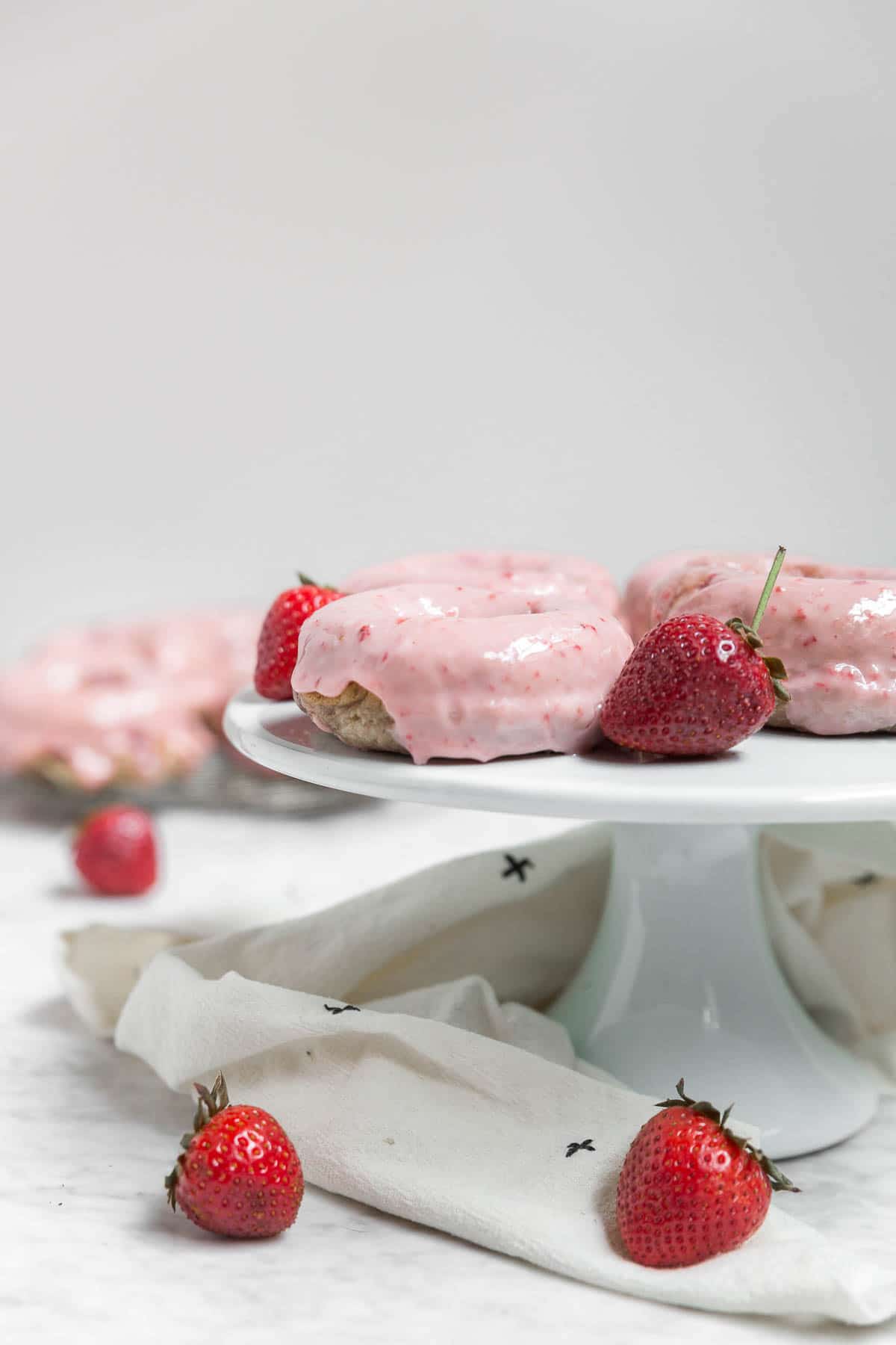A cake stand with pink gluten-free vegan baked strawberry donuts with fresh strawberries and a white linen.