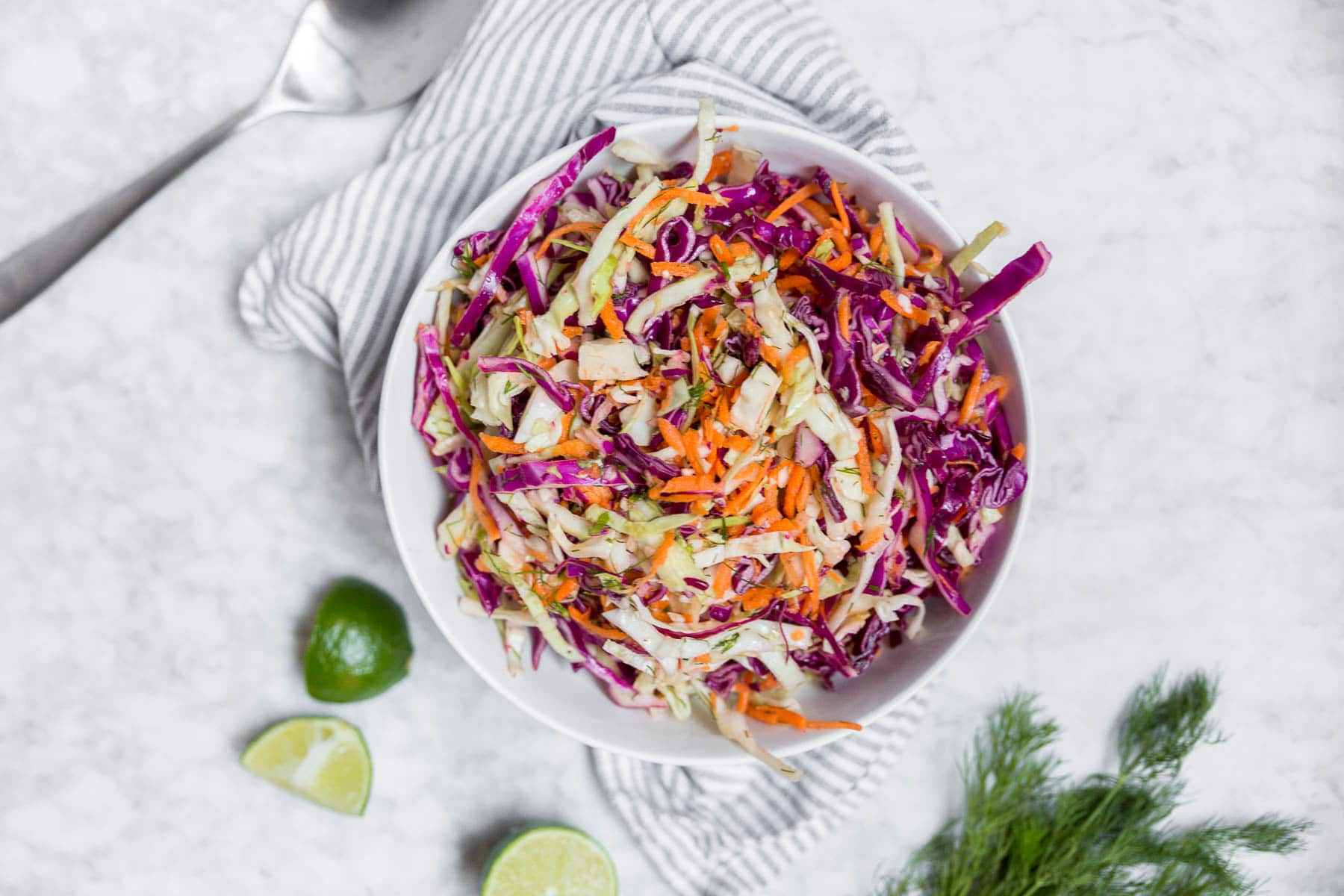 An aerial view of a white bowl on a marble table containing red cabbage carrot dill slaw with lime wedges and fresh dill.