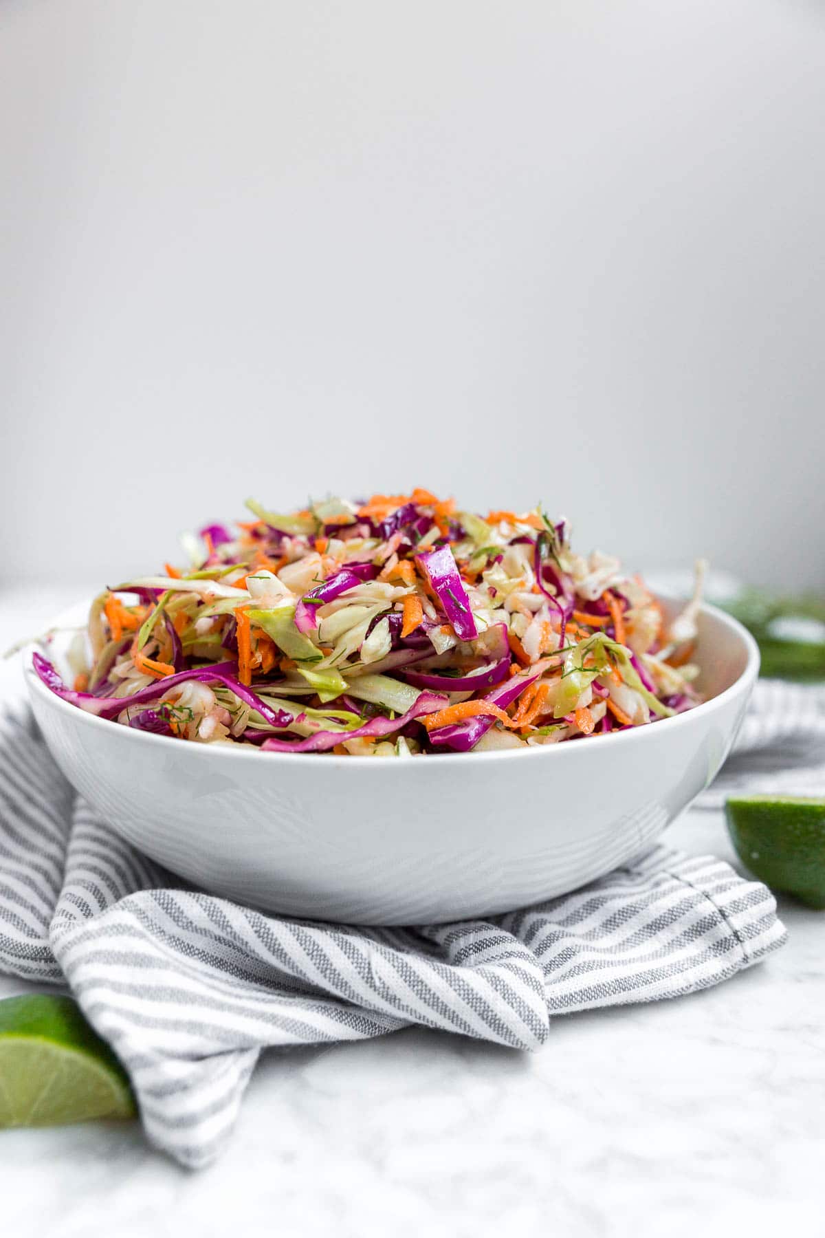 A straight on view of a white bowl containing cabbage carrot coleslaw with dill vinaigrette and lime wedges on a table.