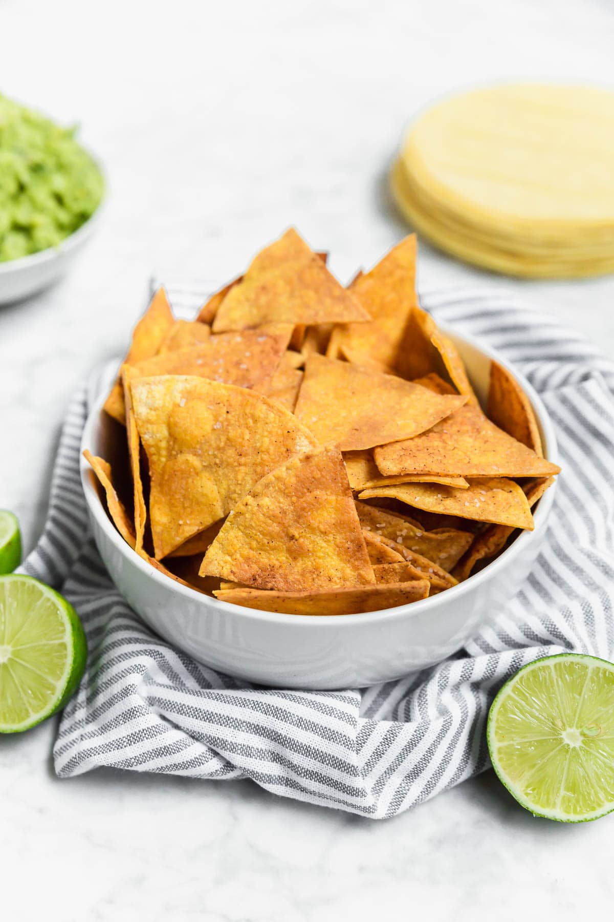 A white bowl filled with golden, crispy Baked Chili Lime Tortilla Chips sits on a striped cloth. Sliced limes and a stack of corn tortillas are nearby, with a bowl of guacamole in the background.