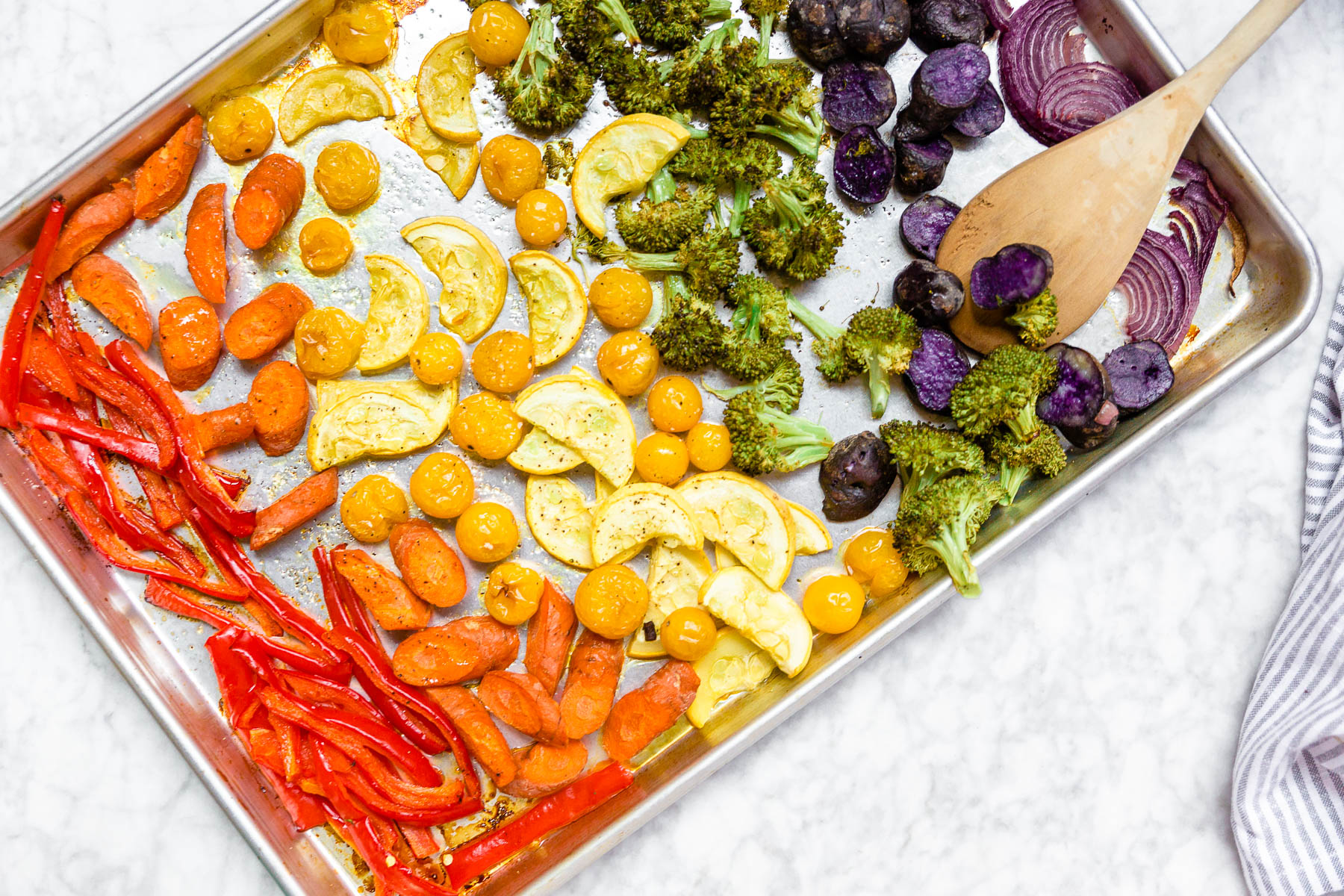 A sheet pan with roasted rainbow vegetables on it including red bell peppers, carrots, yellow squash, yellow tomatoes, broccoli, baby purple potatoes, and red onions with a wooden spoon.