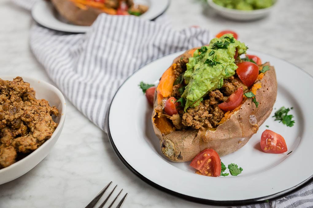 A baked stuffed sweet potato on a white plate, topped with seasoned ground meat, cherry tomatoes, guacamole, and cilantro, sits beside a striped napkin and another plate and bowl of meat in the background.