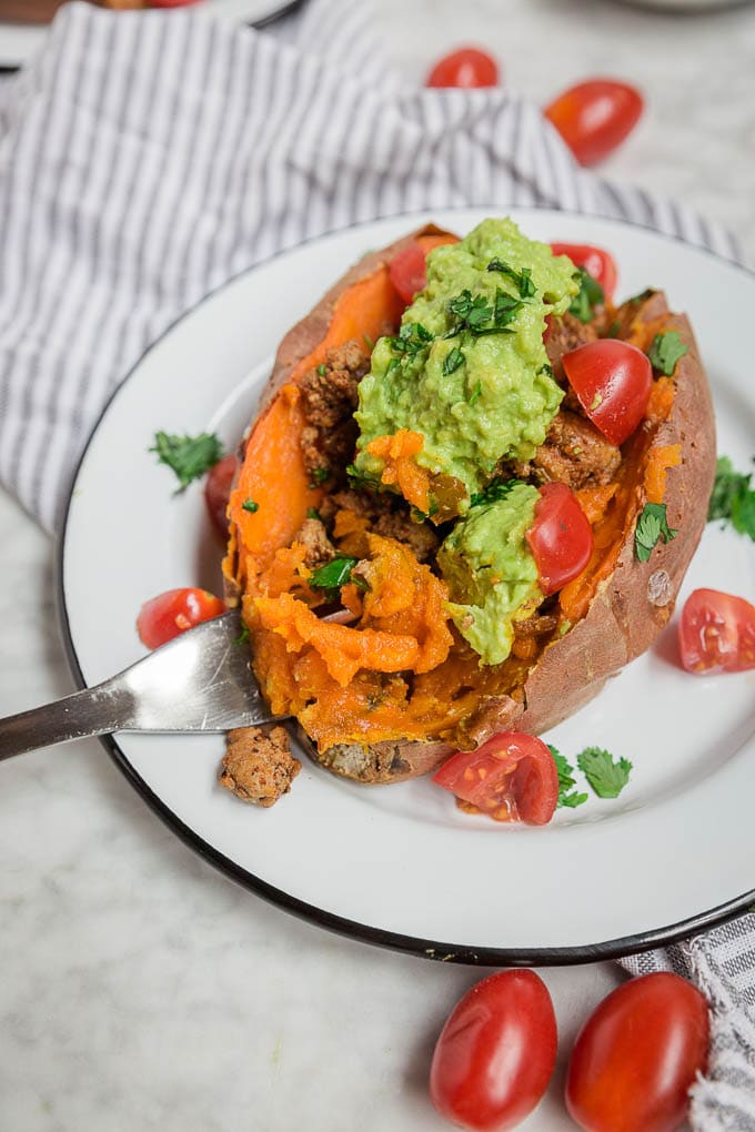 A stuffed sweet potato on a white plate, filled with ground meat and topped with guacamole, chopped cilantro, and halved cherry tomatoes sits beside a fork, with more cherry tomatoes scattered around the plate.