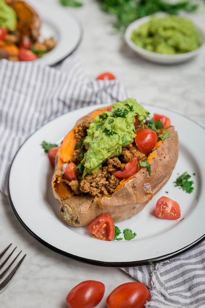 A stuffed sweet potato on a white plate, filled with seasoned ground meat, topped with guacamole and chopped cilantro, and garnished with cherry tomatoes. A striped napkin and extra guacamole sit in the background, highlighting these delicious stuffed sweet potatoes.
