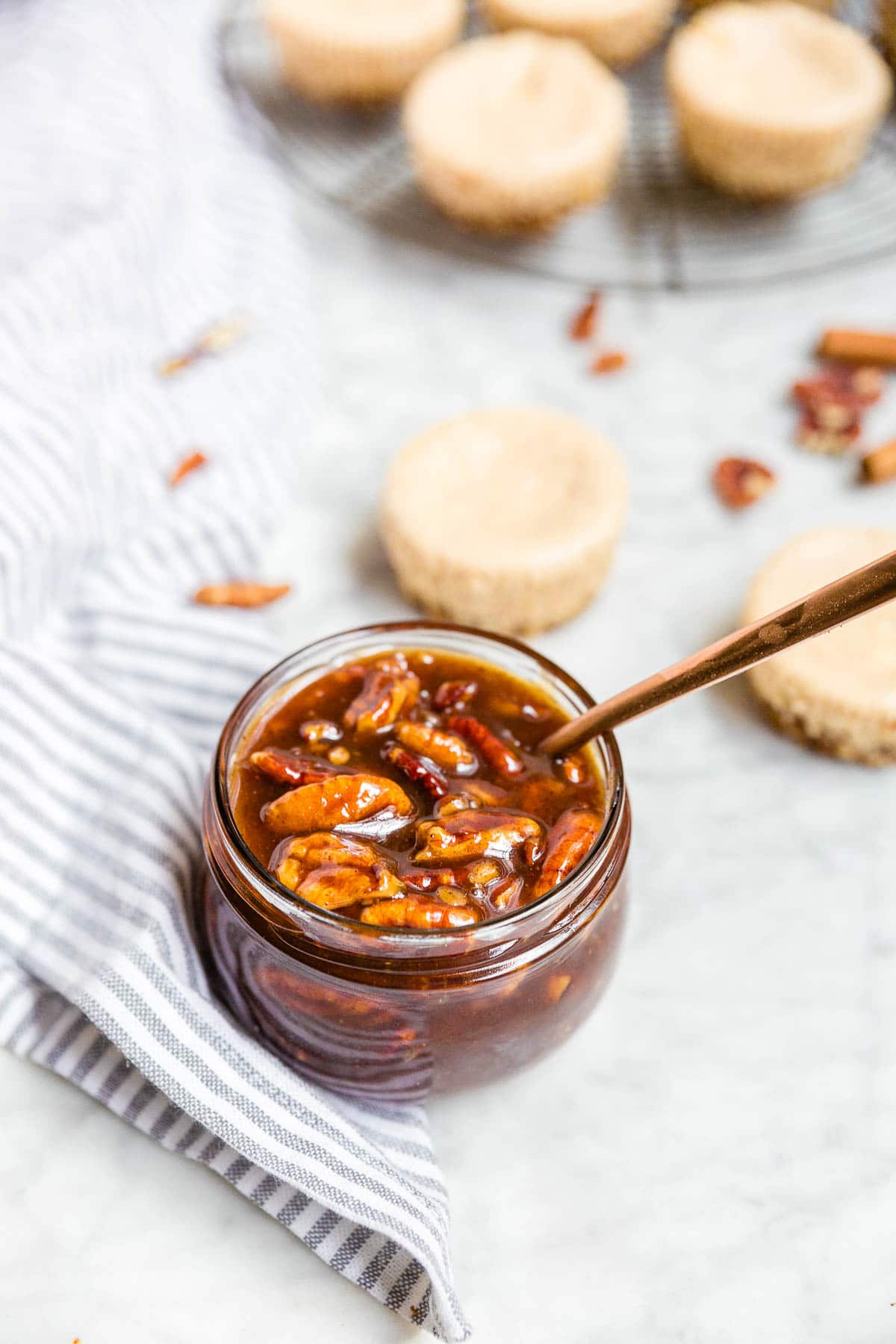 A jar of pecan pie caramel sauce with whole toasted pecans and a spoon ready for drizzling over mini pecan pie cheesecakes.