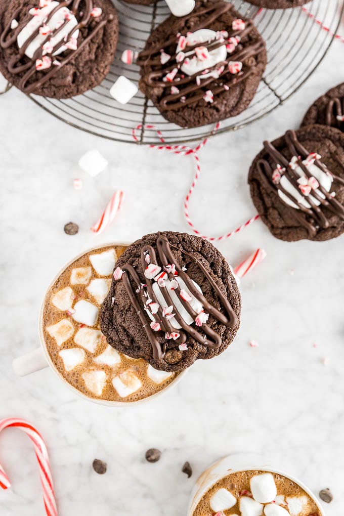 A chocolate peppermint hot chocolate cookie on top of a mug of hot chocolate with mini marshmallows and a baking rack with gluten-free peppermint hot chocolate cookies on it.