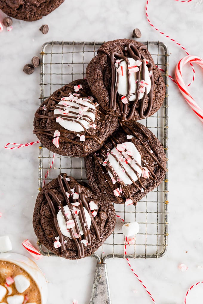 A cooling rack with peppermint hot chocolate cookies topped with marshmallows, a chocolate drizzle and crushed candy canes.