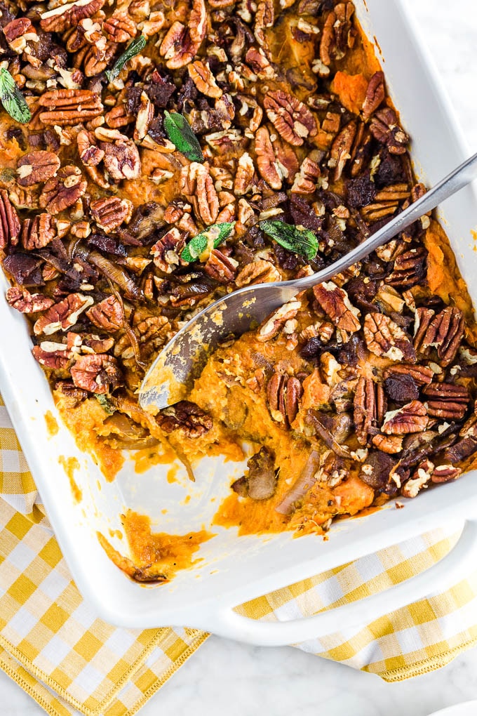 A baking dish with gluten-free savory sweet potato casserole topped with pecans, caramelized onions, bacon, mushrooms and sage with a spoon digging in.