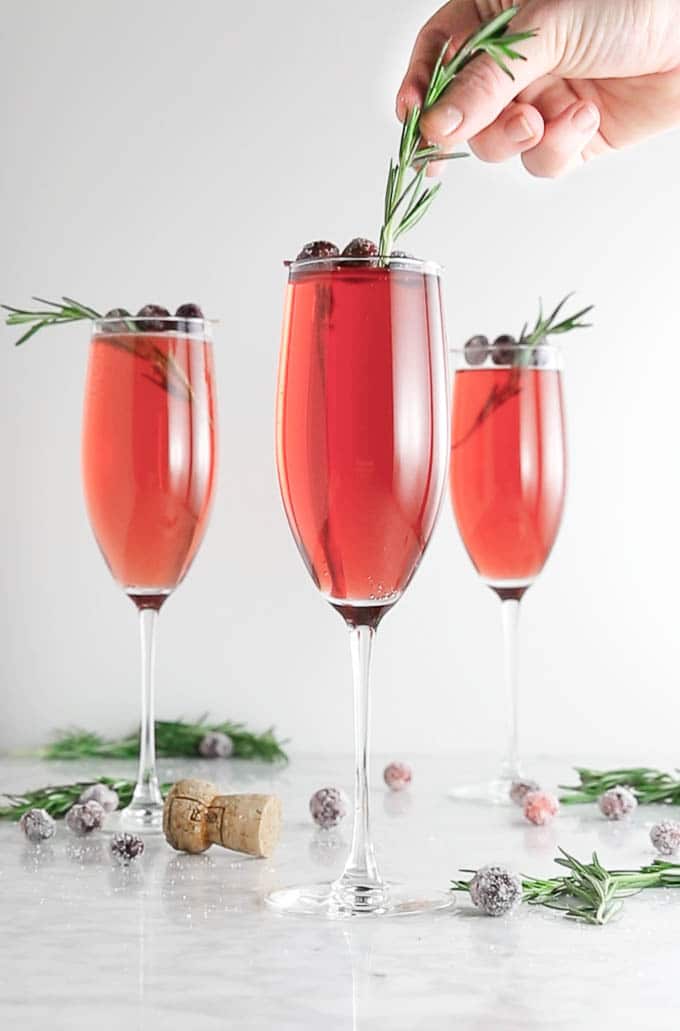 A cranberry mimosa with a sprig of fresh rosemary being placed on top for garnish.