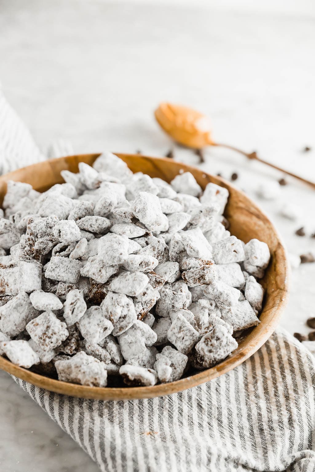 Dairy-free puppy chow in a wooden bowl with a spoonful of peanut butter and chocolate chips and powdered sugar on a linen napkin and table.