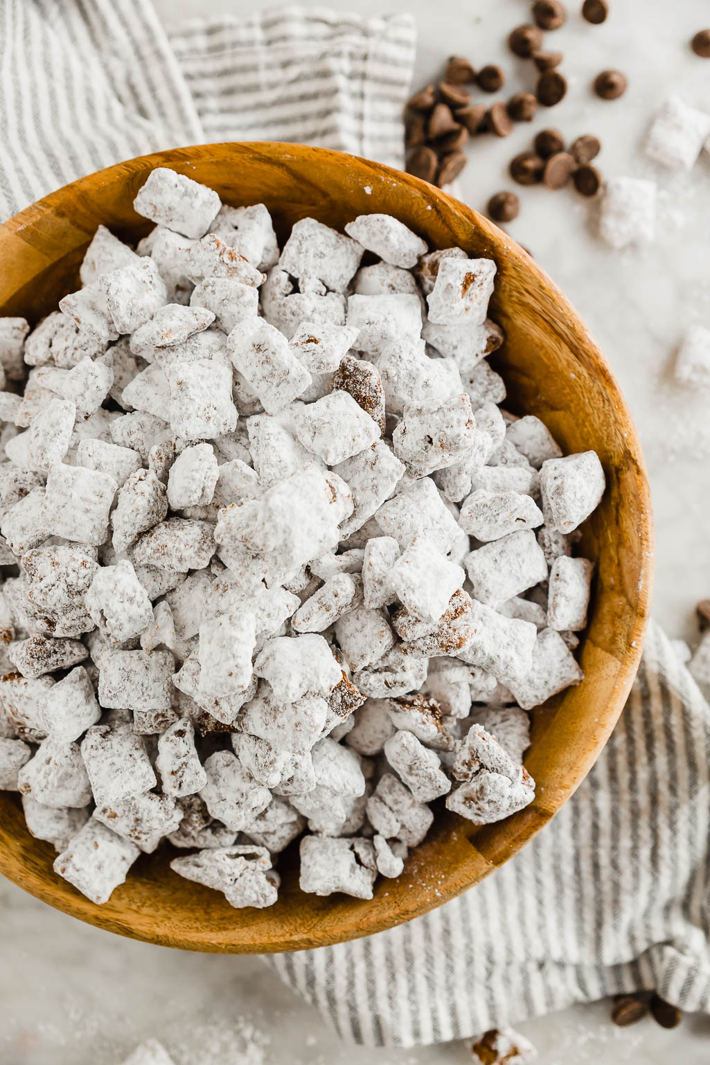Dairy-free puppy chow in a wooden bowl with a spoonful of peanut butter and chocolate chips and powdered sugar on a linen napkin and table.
