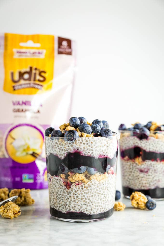 A glass of chia seed pudding layered with Udi's granola, blueberry jam and fresh blueberries with a bag of Udi's Gluten Free Vanilla Granola in the background.