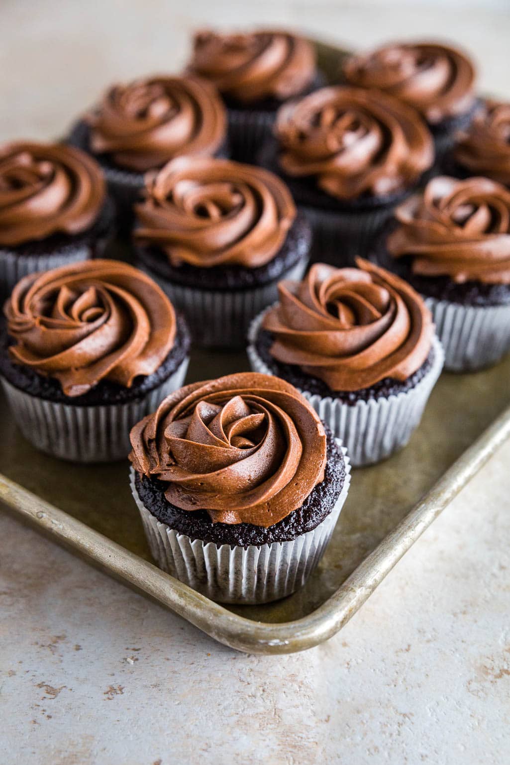A tray of gluten-free chocolate cupcakes with rich, swirled chocolate frosting on top, arranged in neat rows on a metal baking sheet.