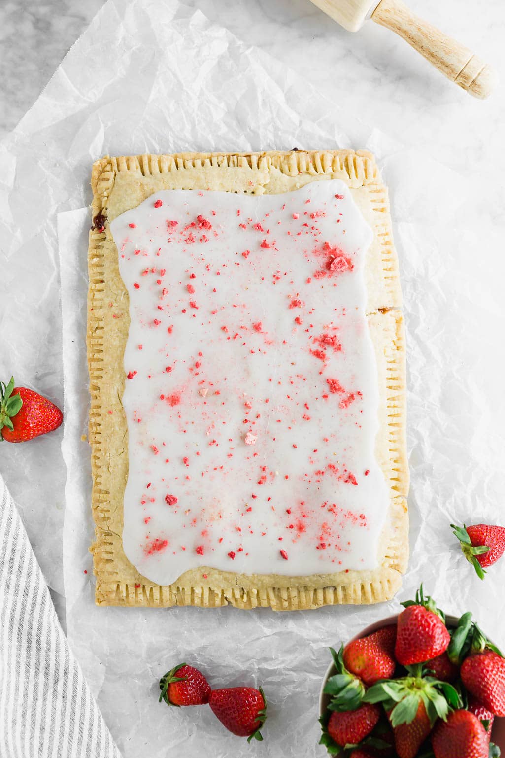 A photo of a giant strawberry pop tart that is gluten free and dairy free on a piece of parchment paper with a bowl of fresh strawberries.