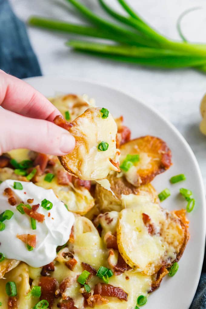 A photo of a plate of potato nachos with yellow potatoes, irish cheddar, green onion, bacon and sour cream with a hand picking a piece of irish nachos up to eat.